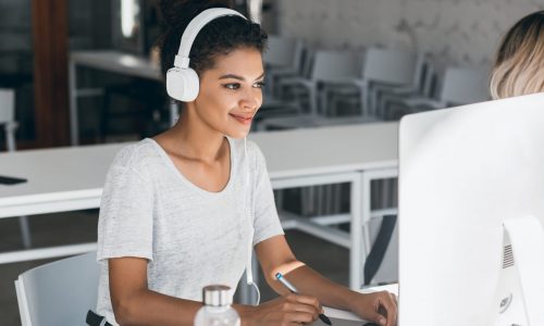 Elegant african web-designer doing her job in office with smile. Attractive black woman in white headphones working in call-center, sitting near computer.